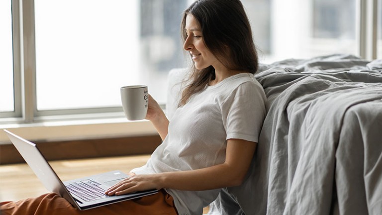 Woman working on a laptop and drinking coffee, sitting on the floor near the bed